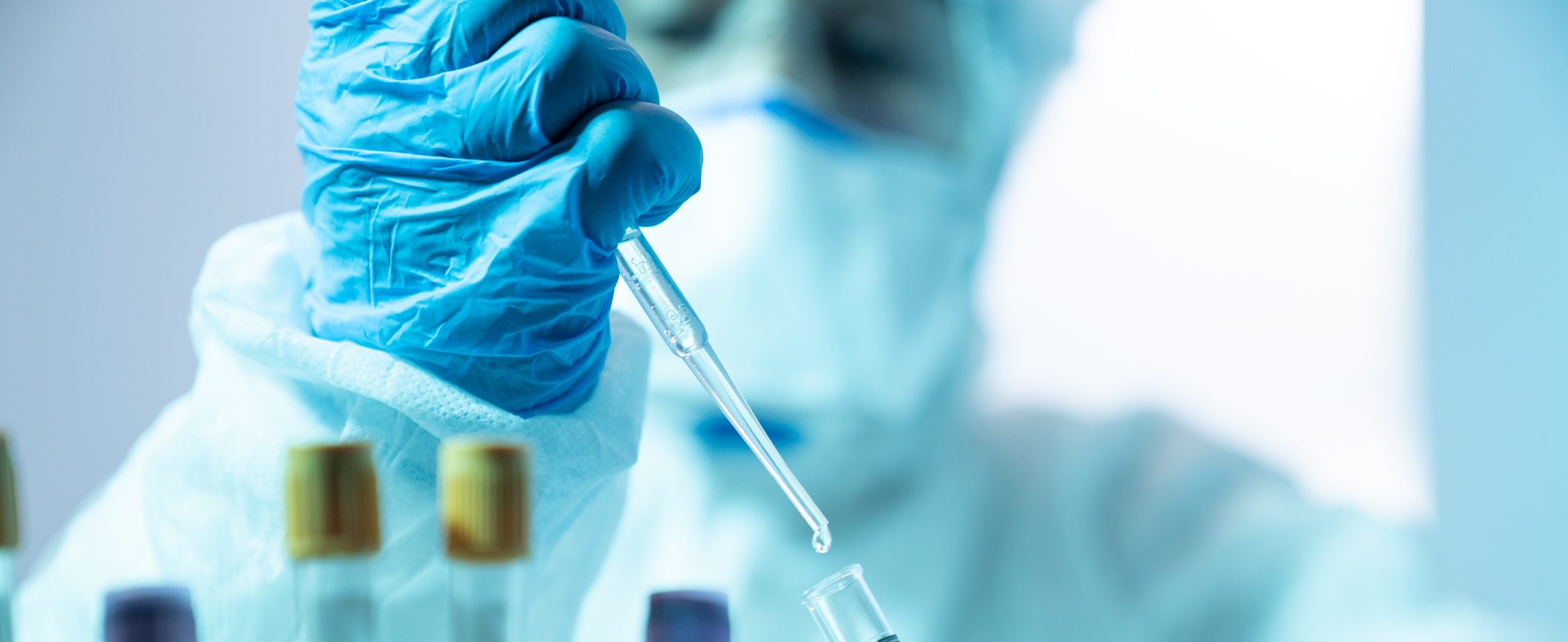 Close up photo of female scientist holding a laboratory pipette and a blood sample tube for covid-19 in laboratory. She is wearing a protective suit, laboratory glasses, surgical mask and surgical gloves. Selective focus on tube. Shot with a full frame mirrorless camera.